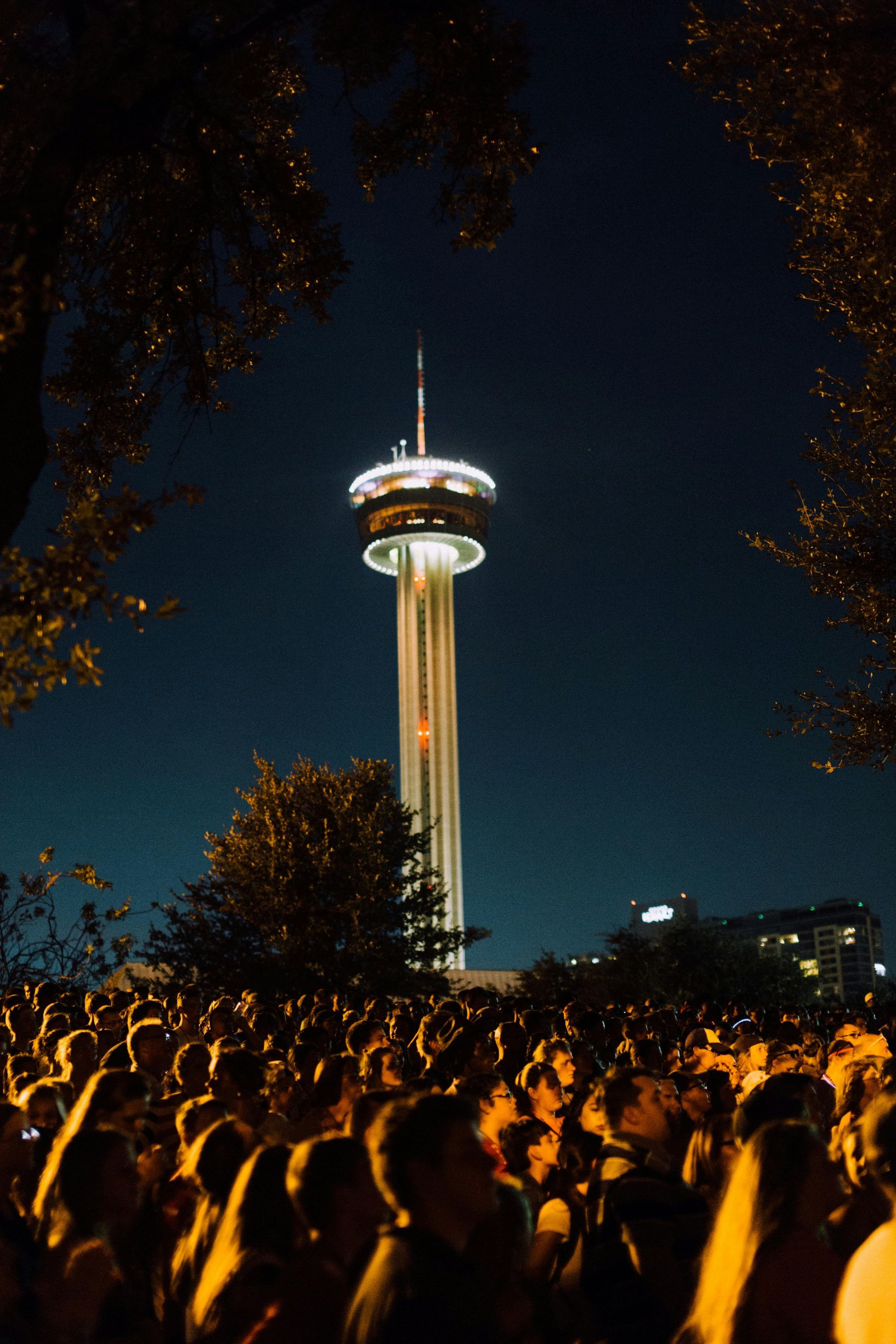 A crowd of people are standing in front of the Tower of the Americas in San Antonio at night.