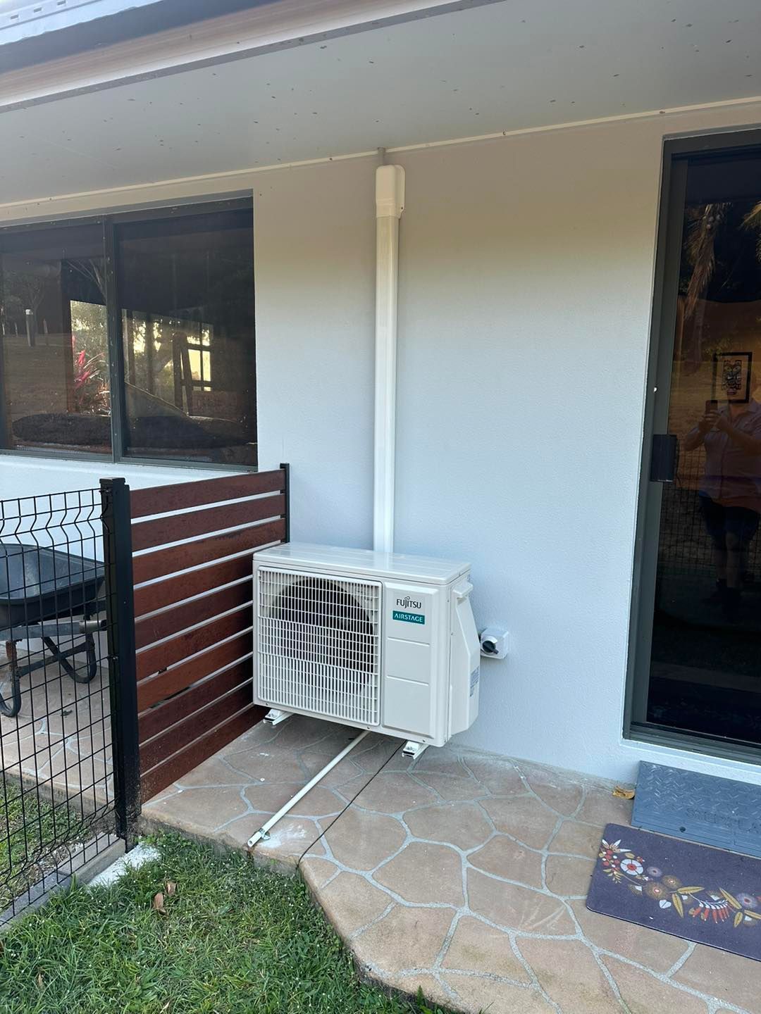 An outdoor air conditioning unit installed on a white wall next to a sliding glass door and a wooden privacy screen.
