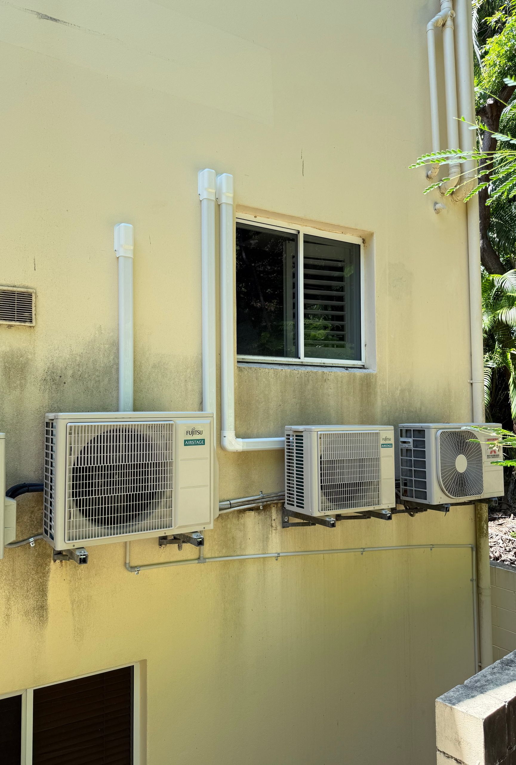Three exterior air conditioning units mounted on a beige building wall below a window.
