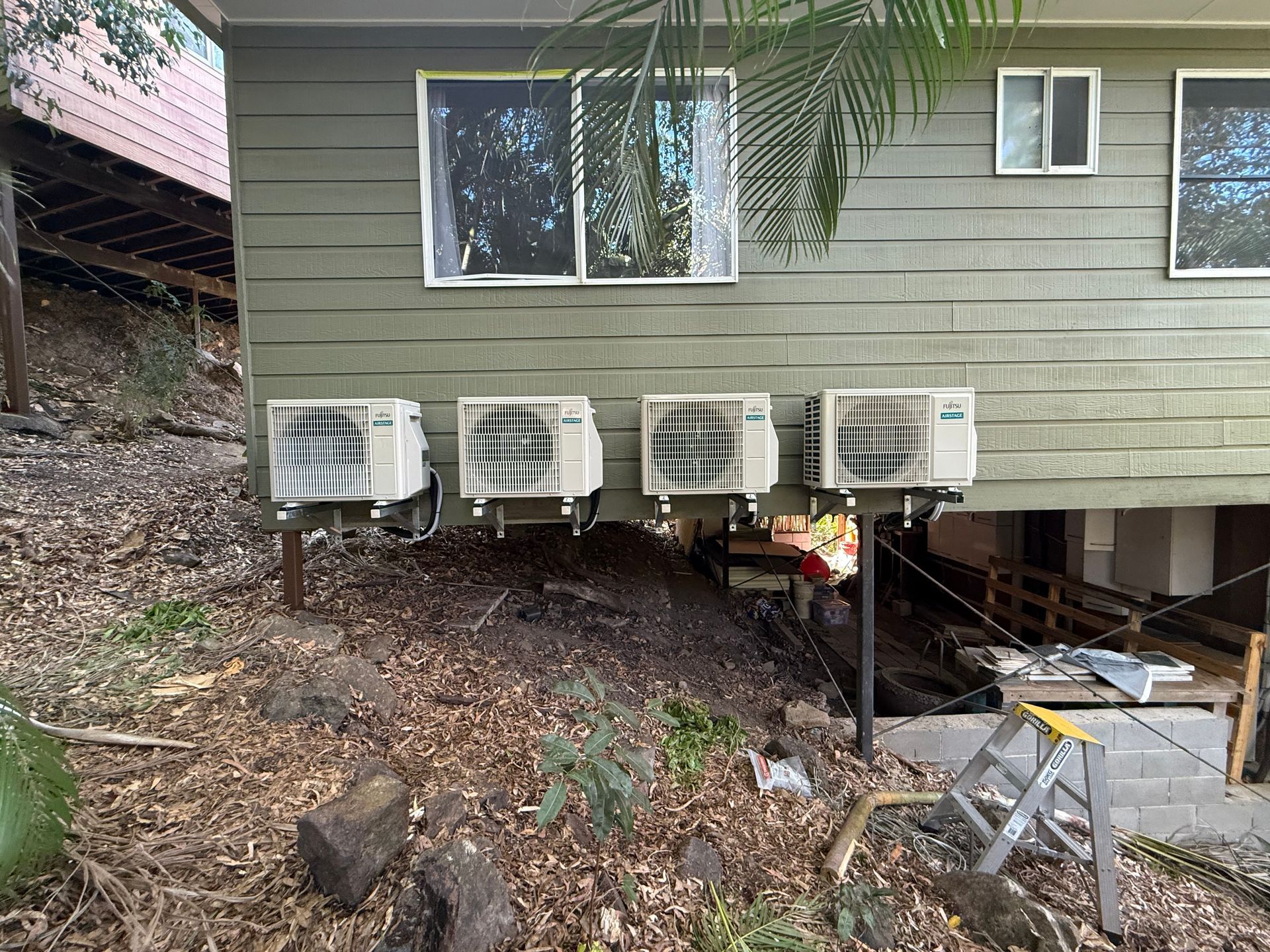 Four air conditioning units mounted on the exterior wall of a green, elevated wooden house above a sloped, wooded yard.