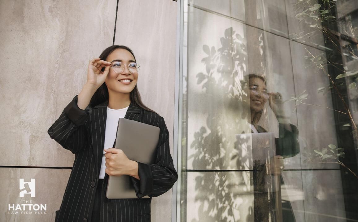 A woman in a suit and glasses is standing in front of a building holding a laptop.