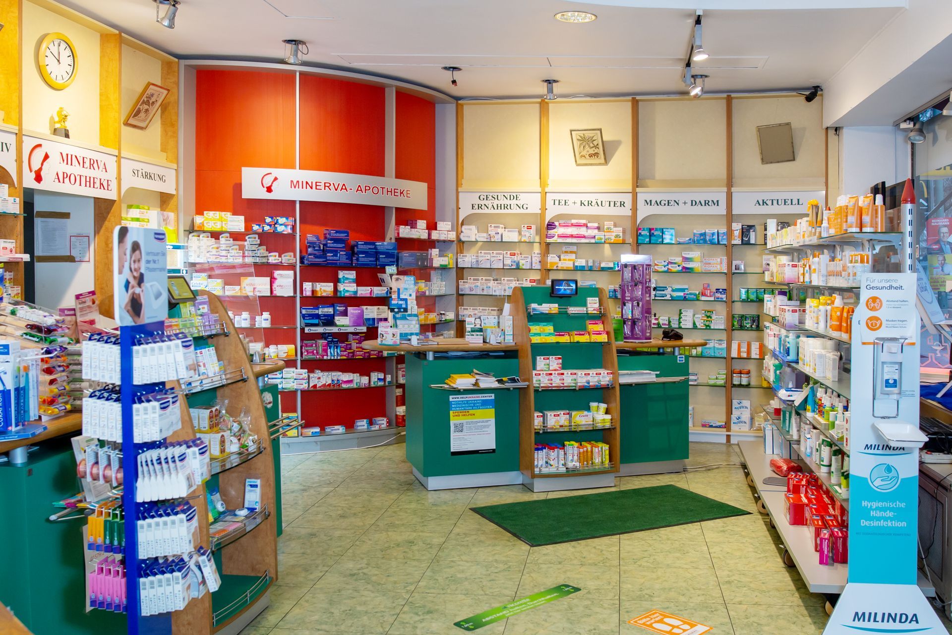 Innenansicht einer hellen Apotheke mit grünen Theken, gefüllten Regalen und roten Akzentwänden.