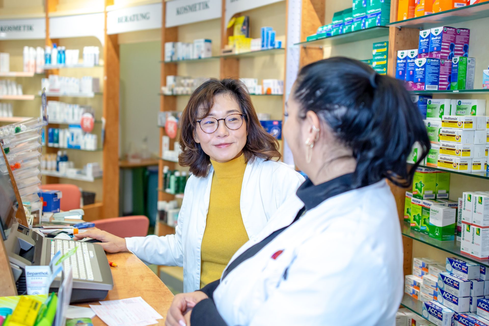 Zwei Apothekenmitarbeiter in weißen Kitteln unterhalten sich, während sie hinter einem mit Medikamenten gefüllten Kassentresen arbeiten.