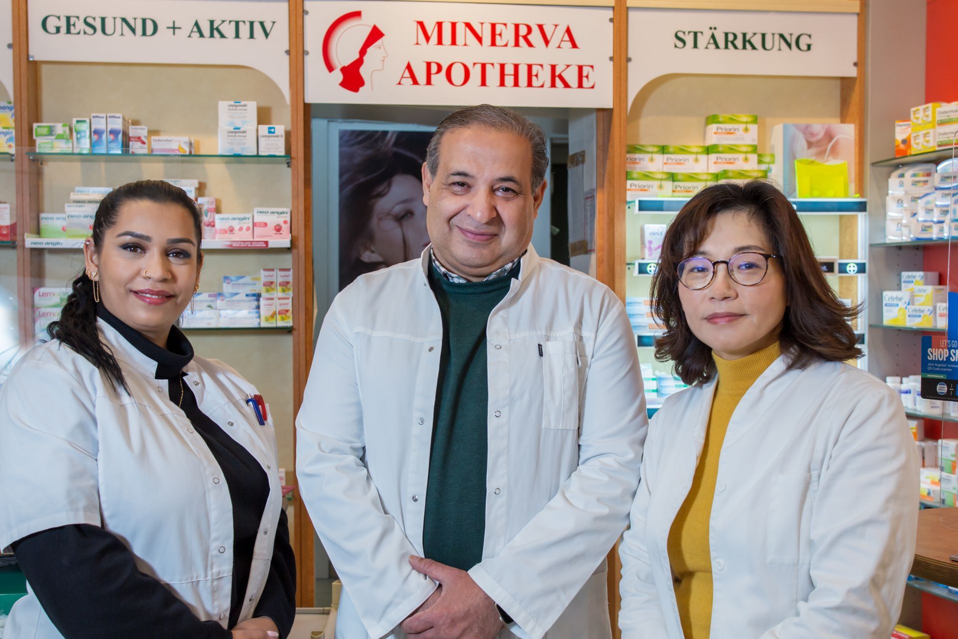 Drei Apothekenmitarbeiter stehen in weißen Laborkitteln in der Minerva Apotheke vor Regalen voller Produkte.