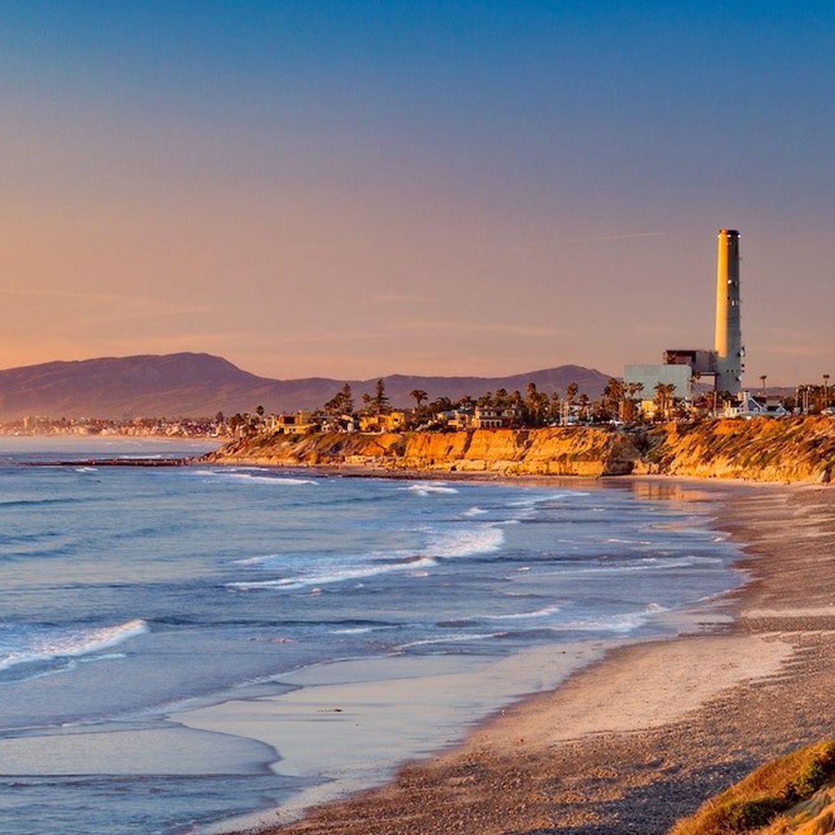 A beach with a power plant in the background at sunset.