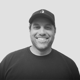 A man wearing a hat and a black shirt is smiling in a black and white photo.