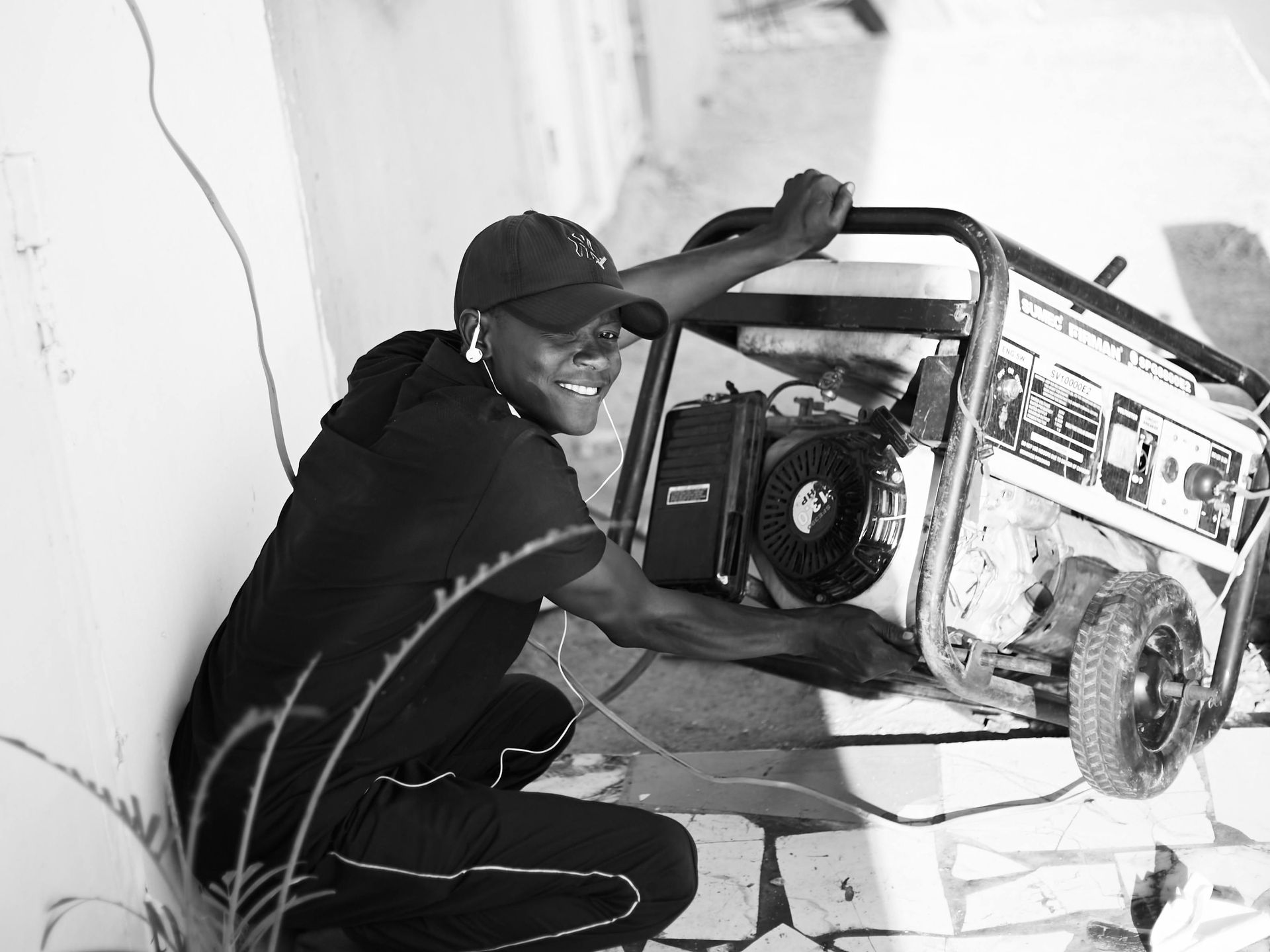Person crouches next to a portable generator, smiling. In an outdoor setting, against a wall. Black and white.