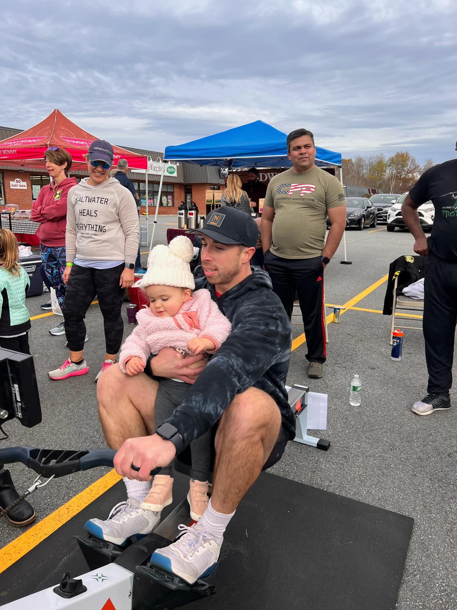 Man rowing machine at outdoor fitness event, holding a baby; colorful tents and onlookers in background