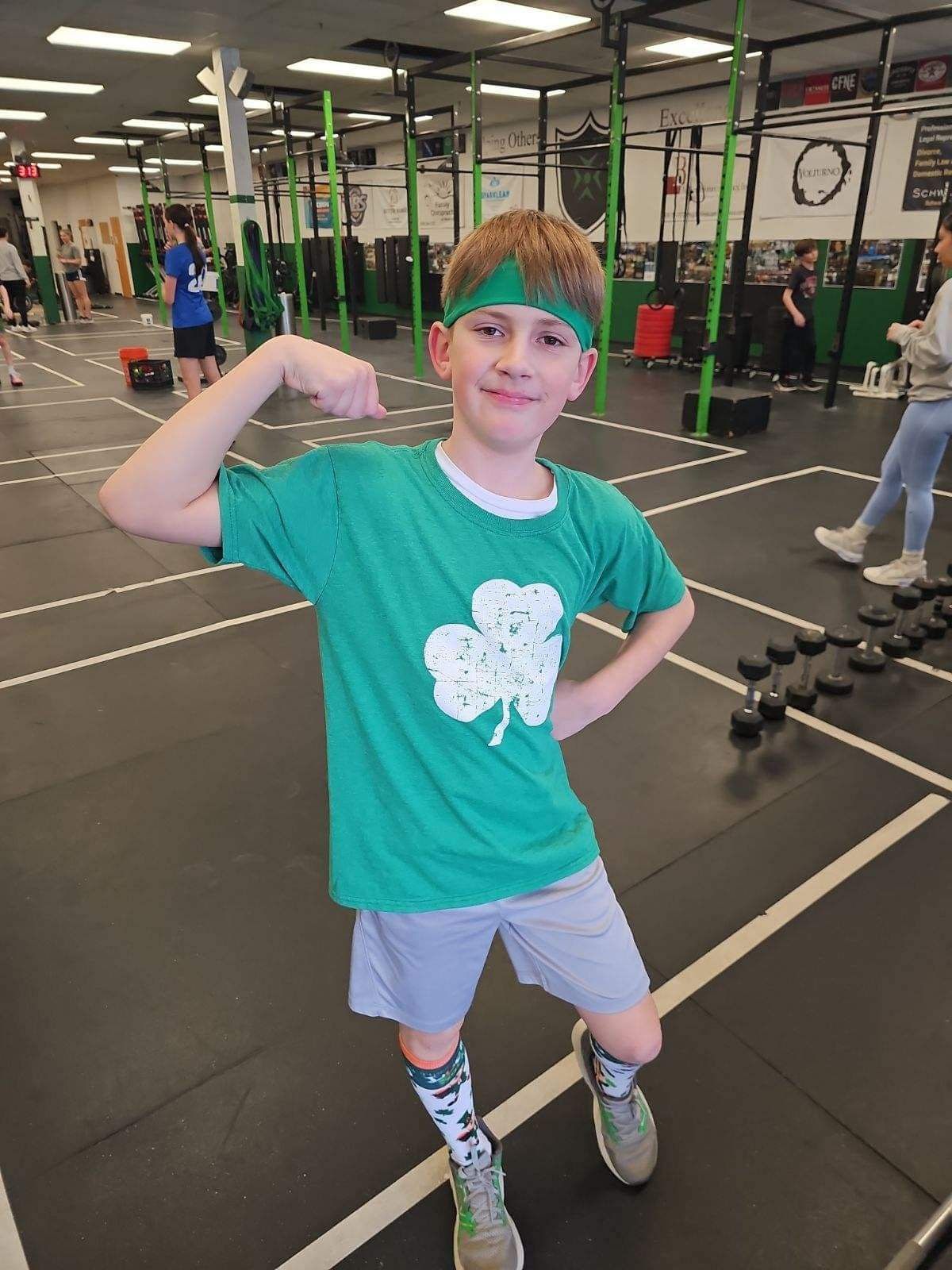 Child flexing in a gym, wearing a teal shirt, gray shorts, and patterned socks.