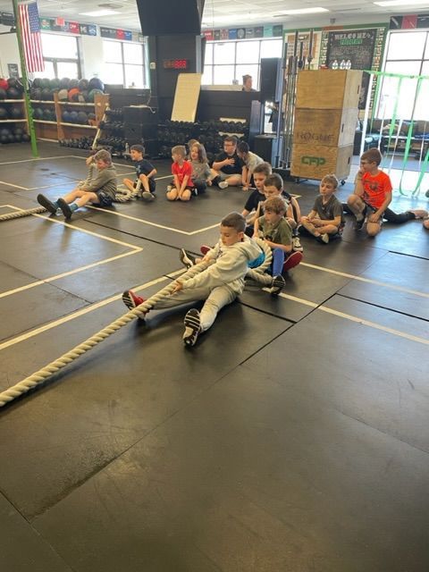 Children sitting on gym floor in a tug-of-war game, holding a thick rope indoors.