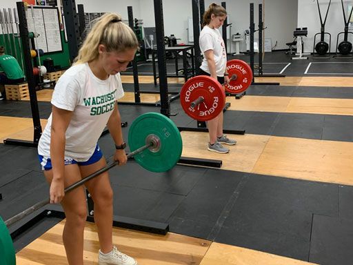Two athletes deadlifting barbells in a gym, with green and red plates on rubber flooring.