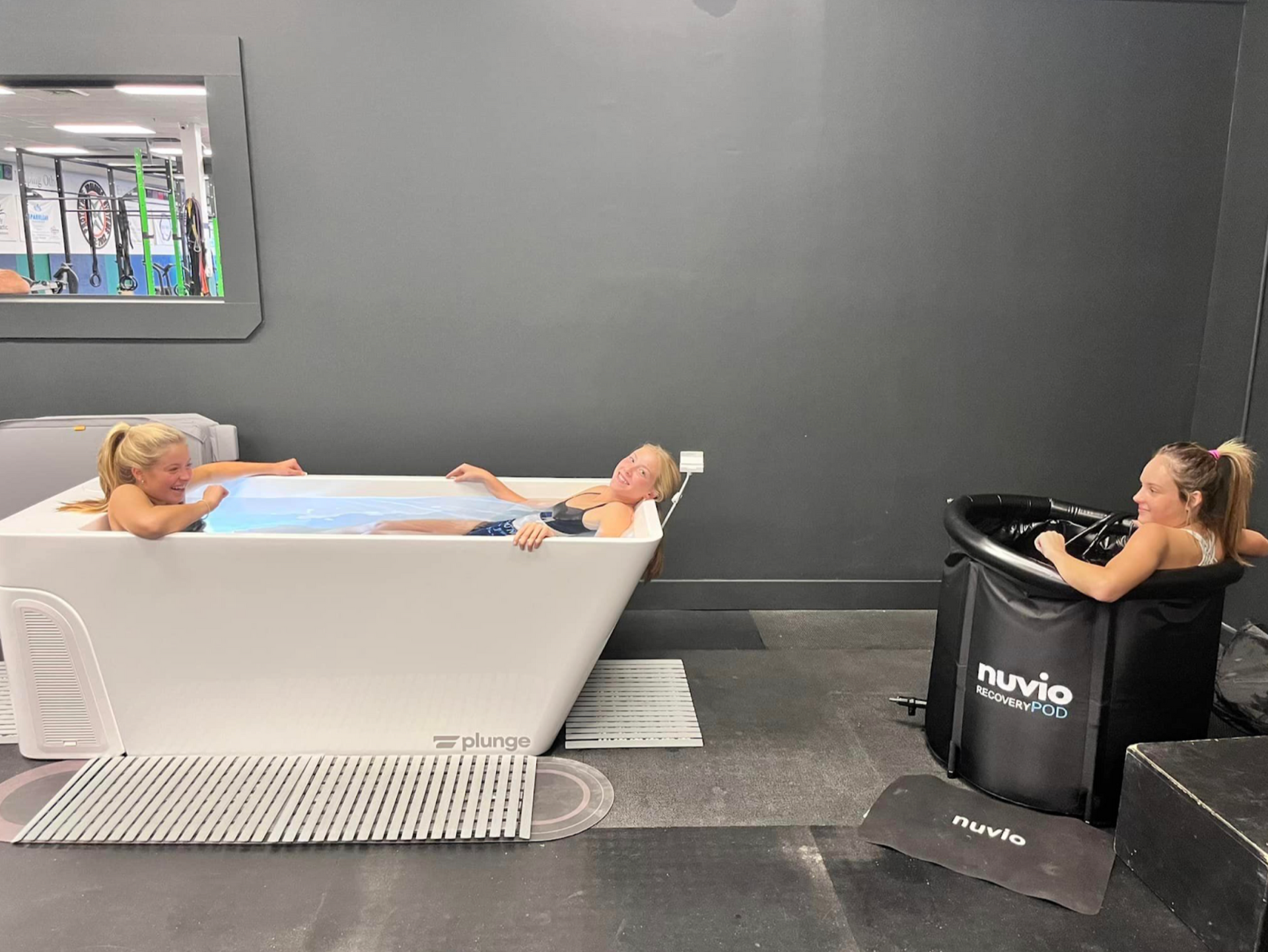 Two people use a bathtub-style bed and a black recovery pod in a gray therapy room.