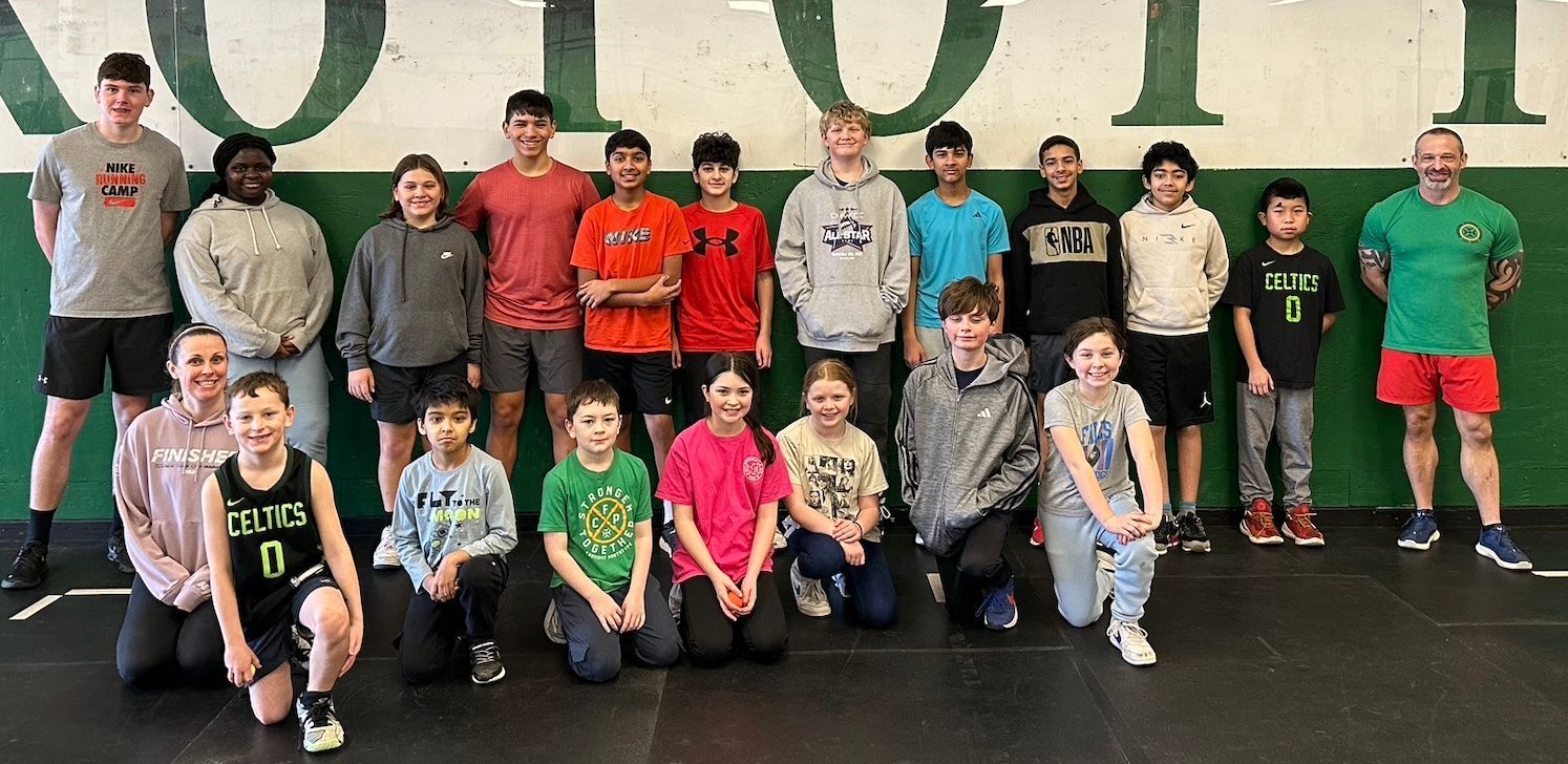 Group of people posing indoors on a gym floor, with a large green wall behind them.