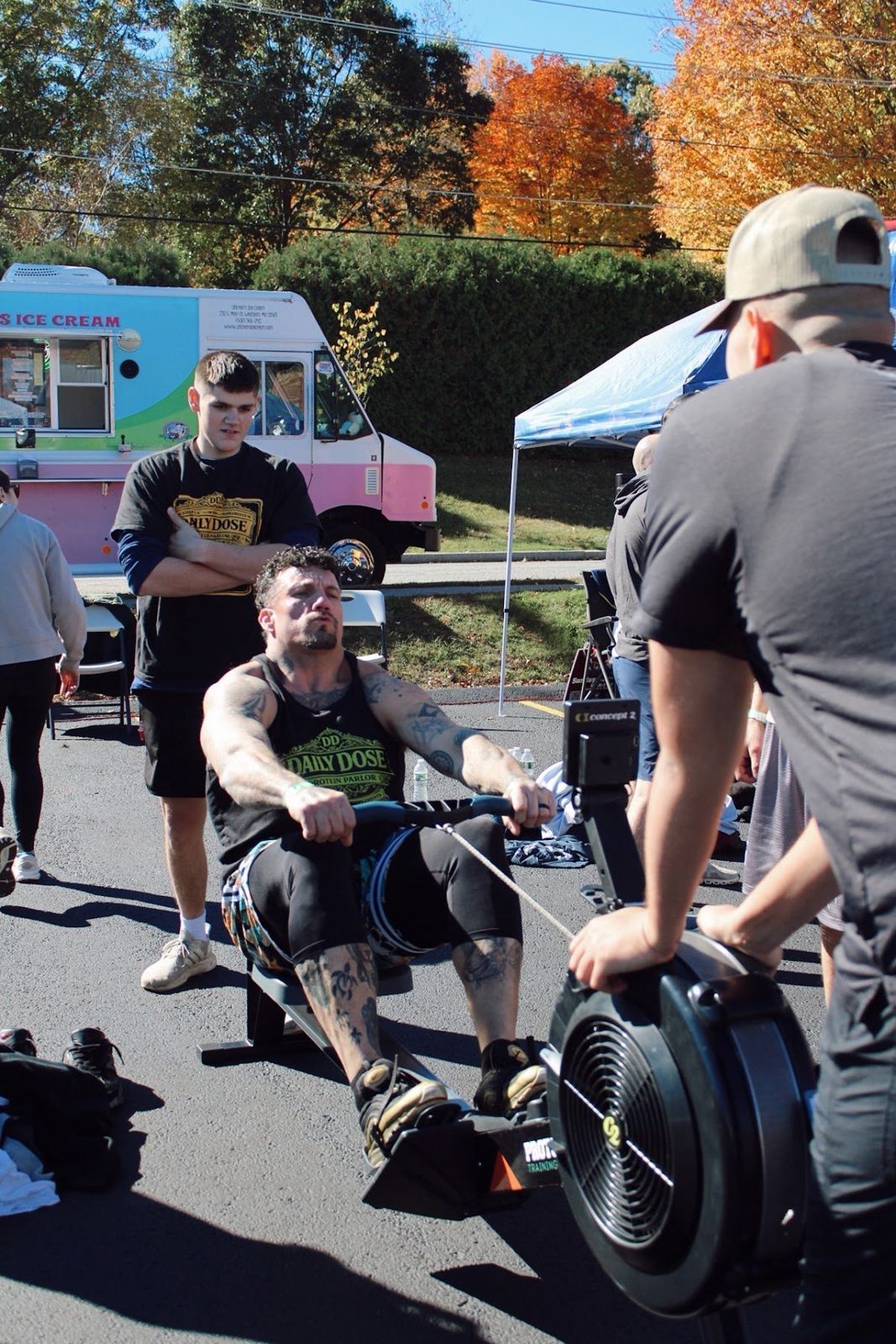 An adult rower in a Daily Dose shirt rowing hard as a teammate coaches him from the front of the erg