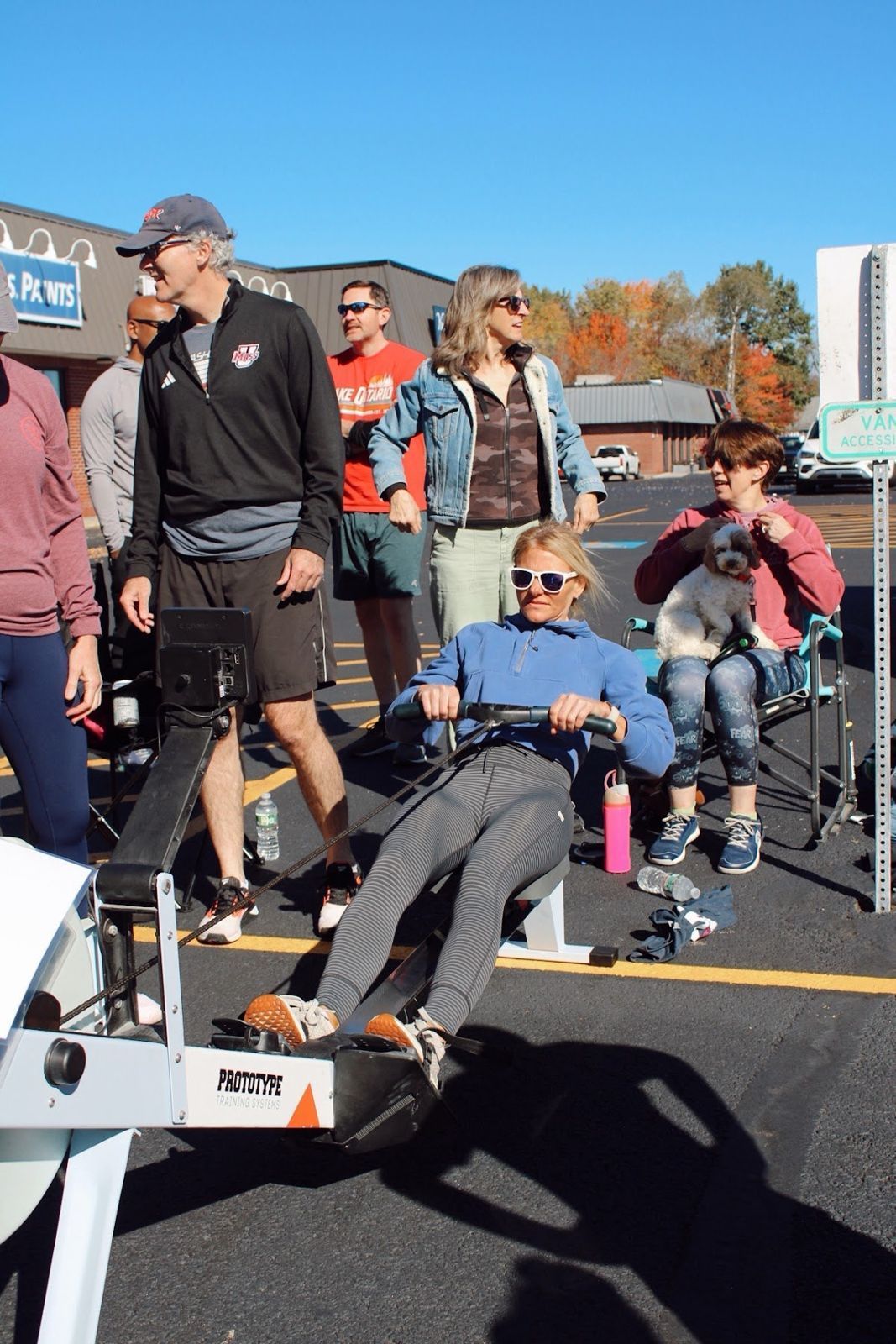 A woman in sunglasses rowing while her crew and a spectator with a small dog watch