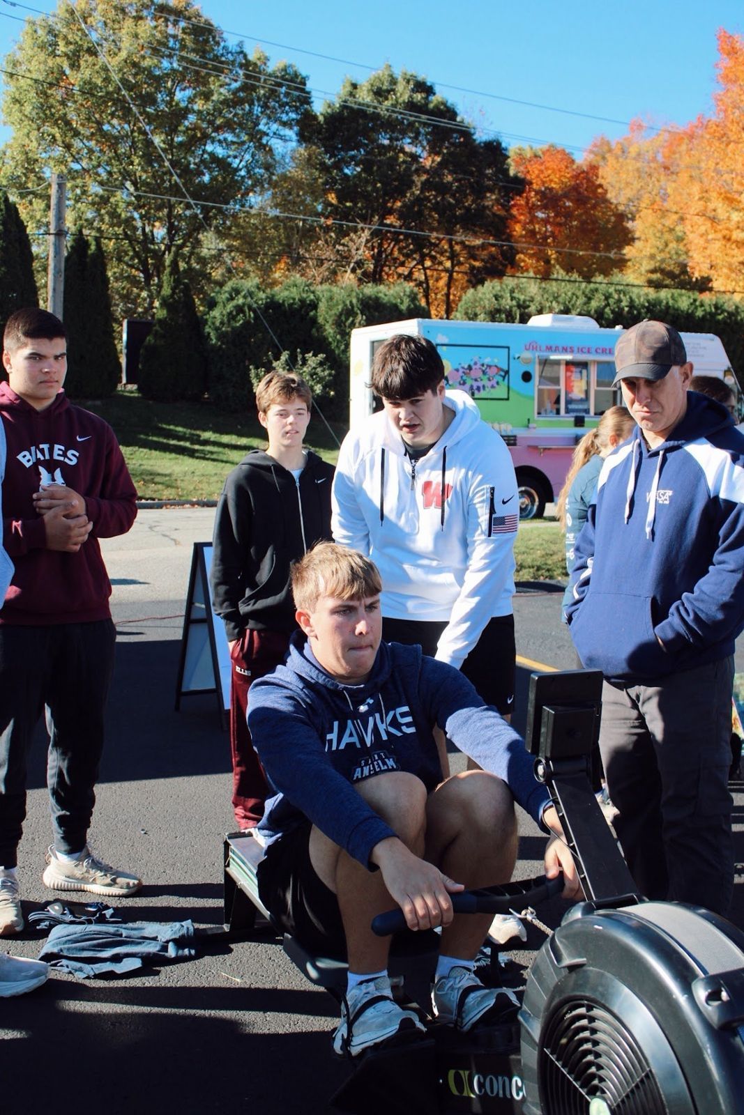 A student rower on a Concept2 erg with his hockey teammates standing behind him