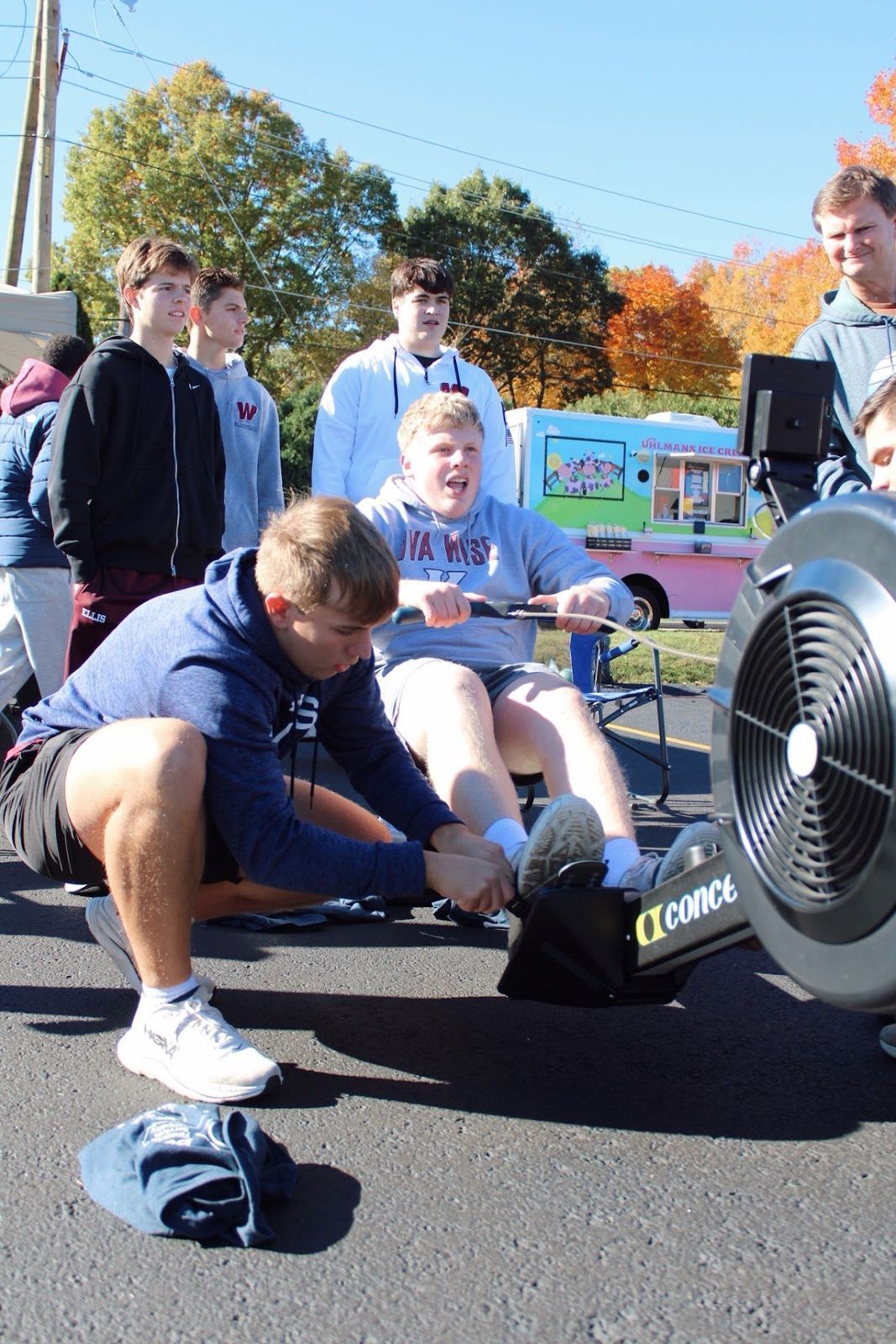 A student rower pulling hard while a teammate kneels and holds his feet on the rowing machine