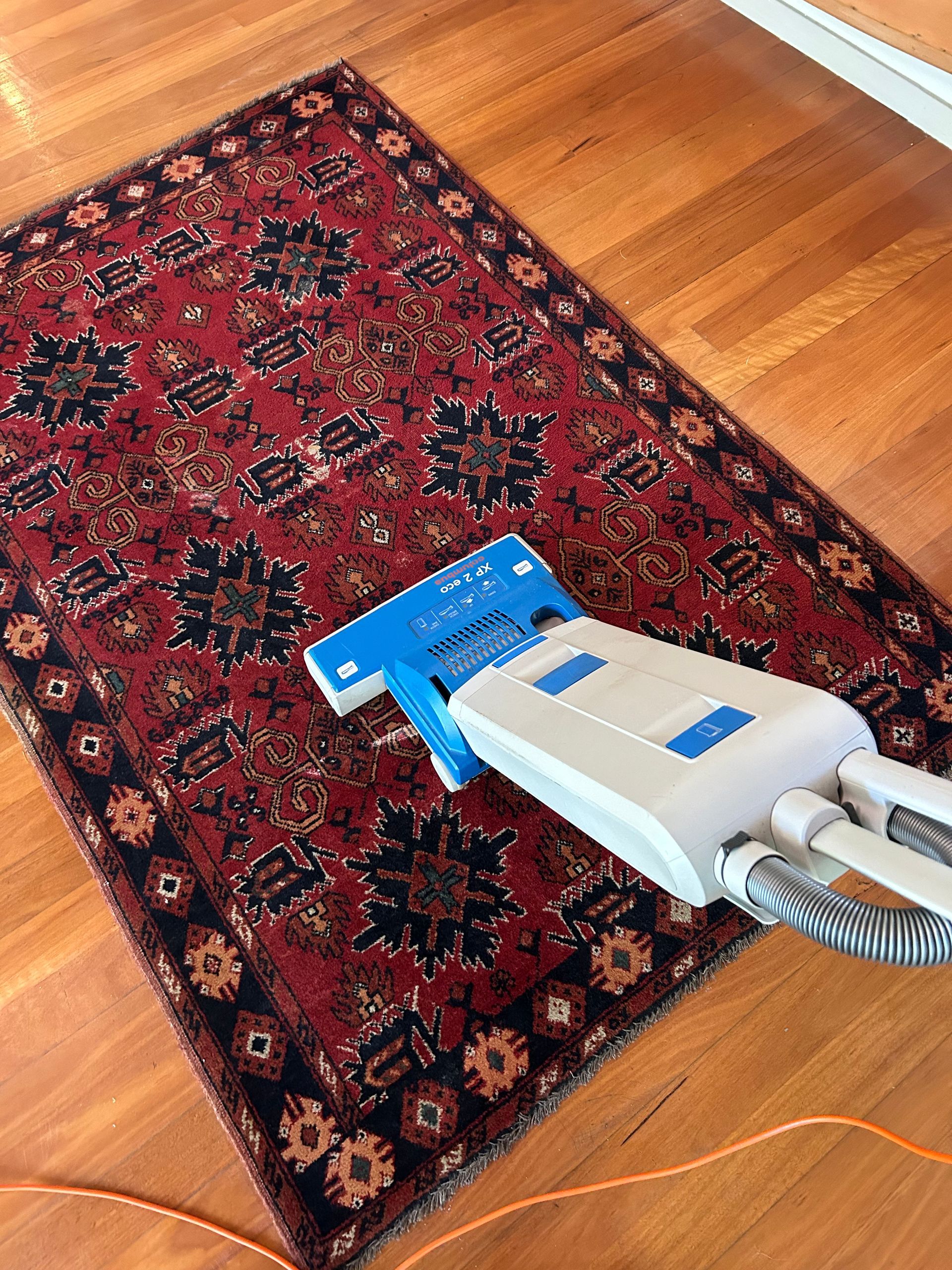 Vacuum cleaner head cleaning a red patterned rug on a hardwood floor — Rugrats Carpet Cleaning Service In Old Bar, NSW