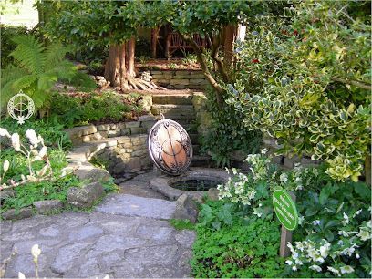 Stone garden with steps, well, and hanging orb. Green foliage surrounds the path; a tree is in the background.