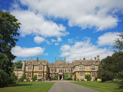 Large stone manor house under a blue sky with fluffy white clouds; a long driveway leads to the entrance.