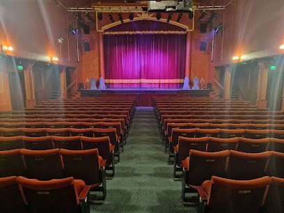 Empty theater auditorium with rows of red seats facing a stage with red curtains lit in purple.