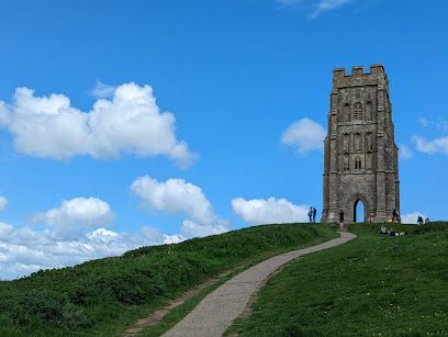 Stone tower on a grassy hill with a path leading up to it, under a blue sky with fluffy clouds.