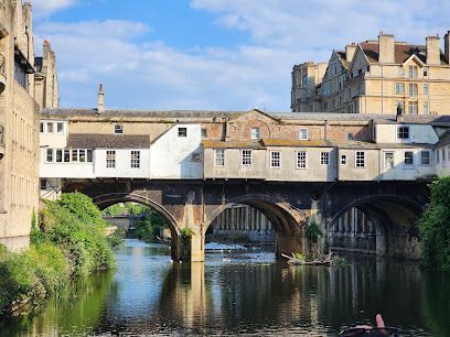 Pulteney Bridge in Bath, England, with shops built along it, over a river. Stone arches, blue sky.