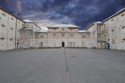 Exterior of an old stone prison with a large empty courtyard under a dramatic, cloudy sky.