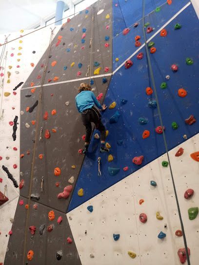 A person climbing an indoor rock wall with colorful holds. Wall sections are gray, white, and blue.