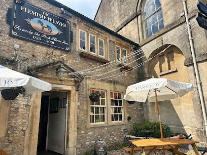 The Flemish Weaver pub exterior with outdoor seating, stone building, sign with horses, sunlight.