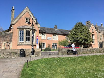Peach-colored building with arched windows, green lawn, person on bench, blue sky.