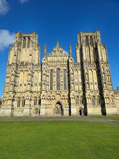 Wells Cathedral exterior against a blue sky. Light stone facade with towers and arched doorway.