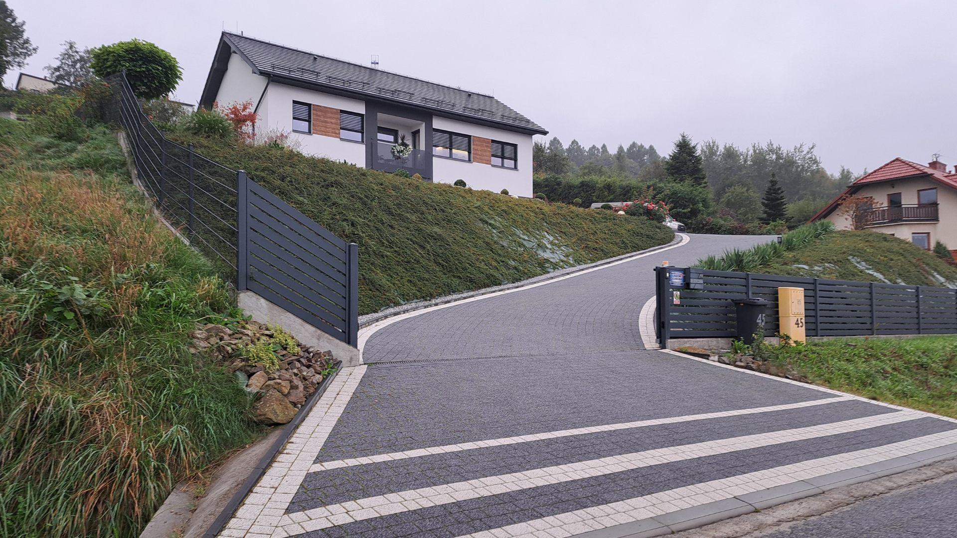 House with gray roof and driveway. Sloping hill covered in greenery, dark gray and white pavers.