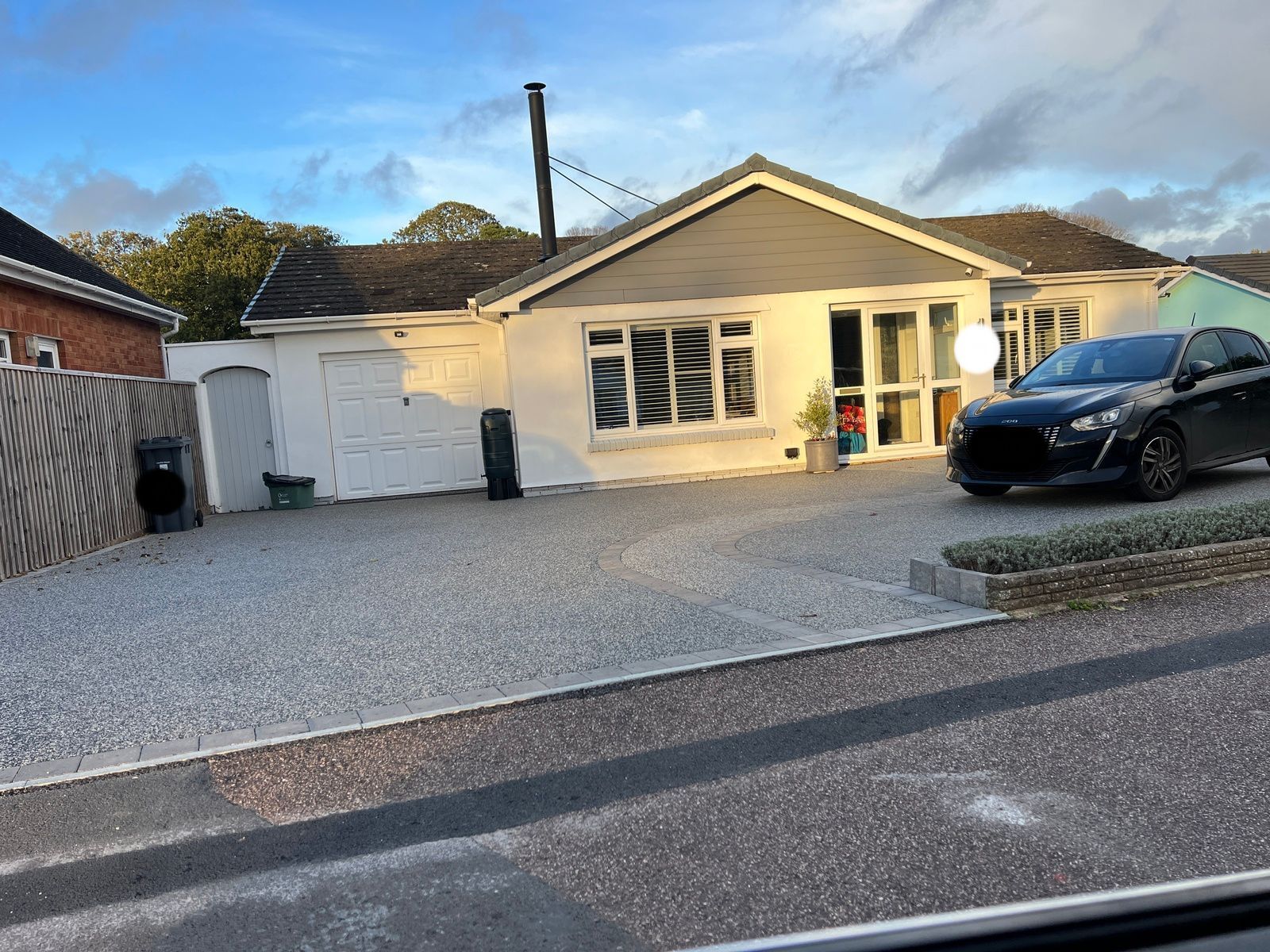 A single-story white house with a gray roof and a black car parked in the driveway. Gravel covers the yard.
