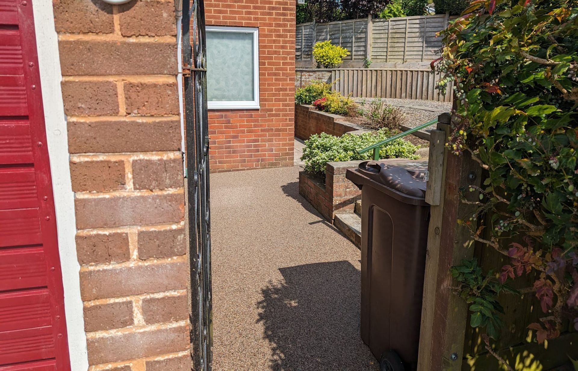 View through a gate: gravel path, brown trash can, brick building, and terraced garden with greenery and a fence.