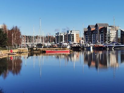 Boats docked at a marina with modern buildings in the background, clear blue sky, water reflections.