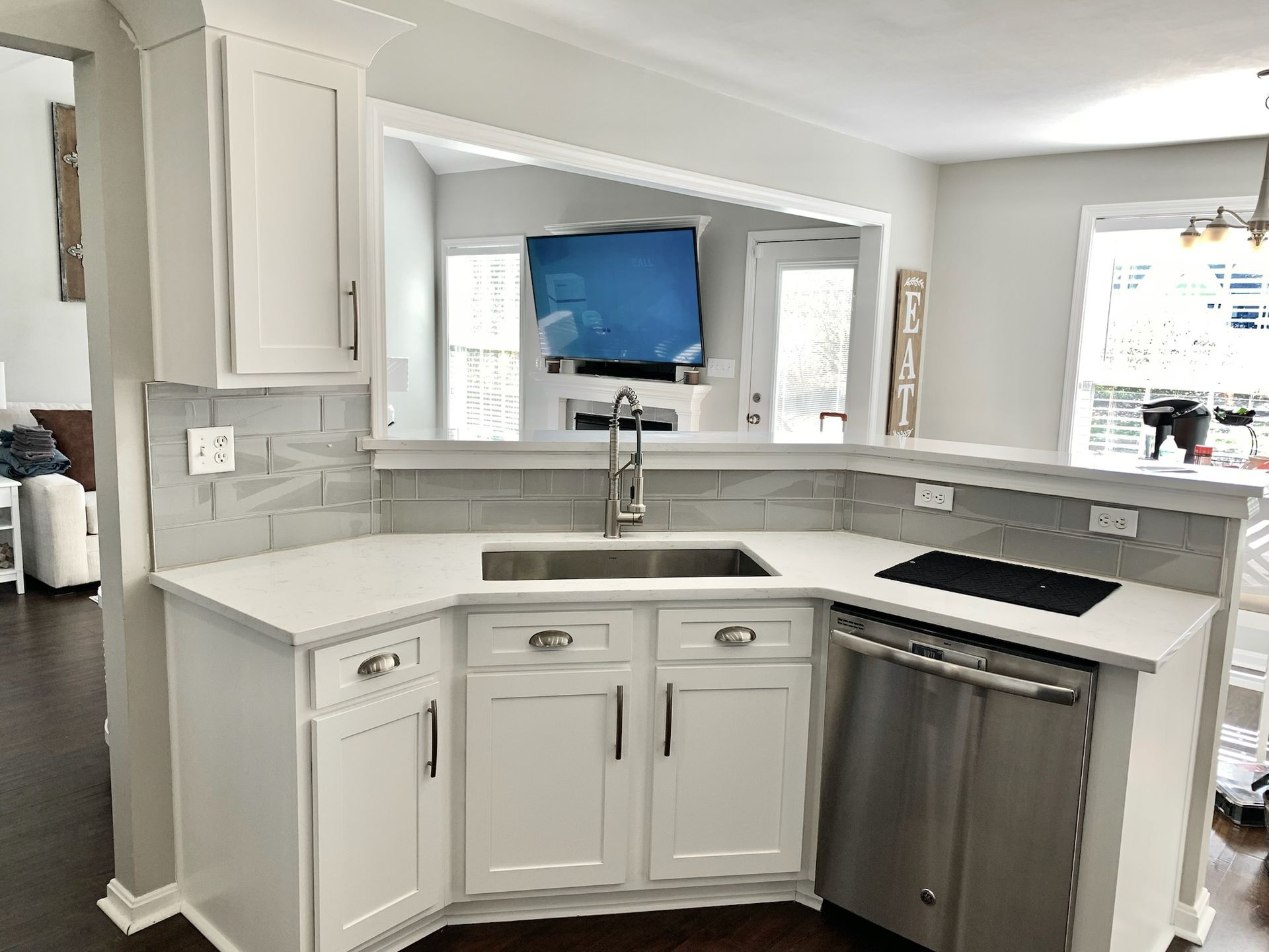 White kitchen with sink and dishwasher; countertop with grey tiled backsplash; opens to a room with a TV.