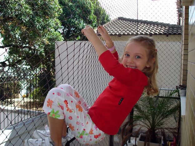 Criança de camisa vermelha e calça florida brincando em um balanço de rede ao ar livre, sorrindo.