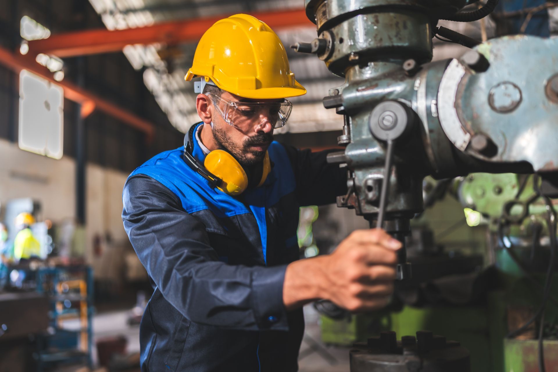 Worker in a yellow hard hat and safety glasses operating a drill press in a factory.