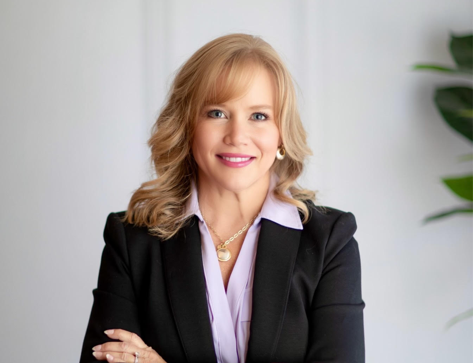 Smiling professional in a black blazer and light blouse, standing with arms crossed against a plain wall.