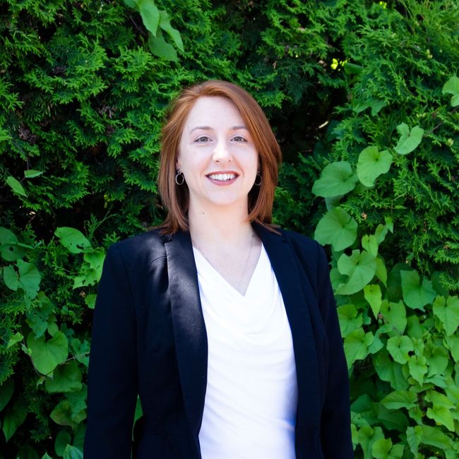 Smiling person in a black blazer and white top standing in front of lush green foliage