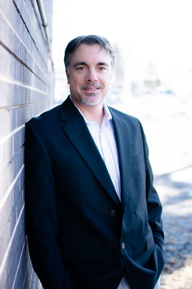 Man in a dark blazer leaning against a wall outdoors, smiling in bright light.