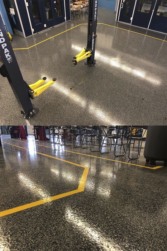 Un piso de concreto con líneas amarillas que delimitan áreas; un elevador de vehículos en un área, mesas y sillas en otra.