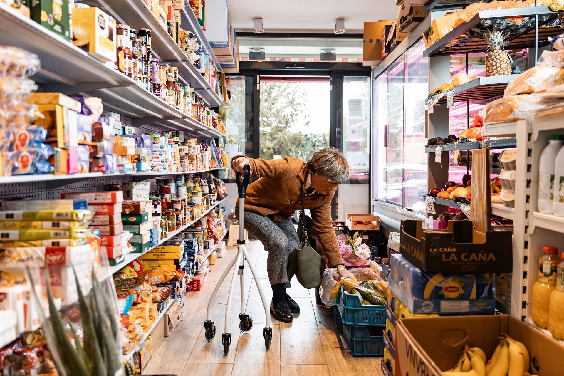 Person stocking shelves in a narrow grocery aisle, with products lining both sides and a refrigerated case ahead.