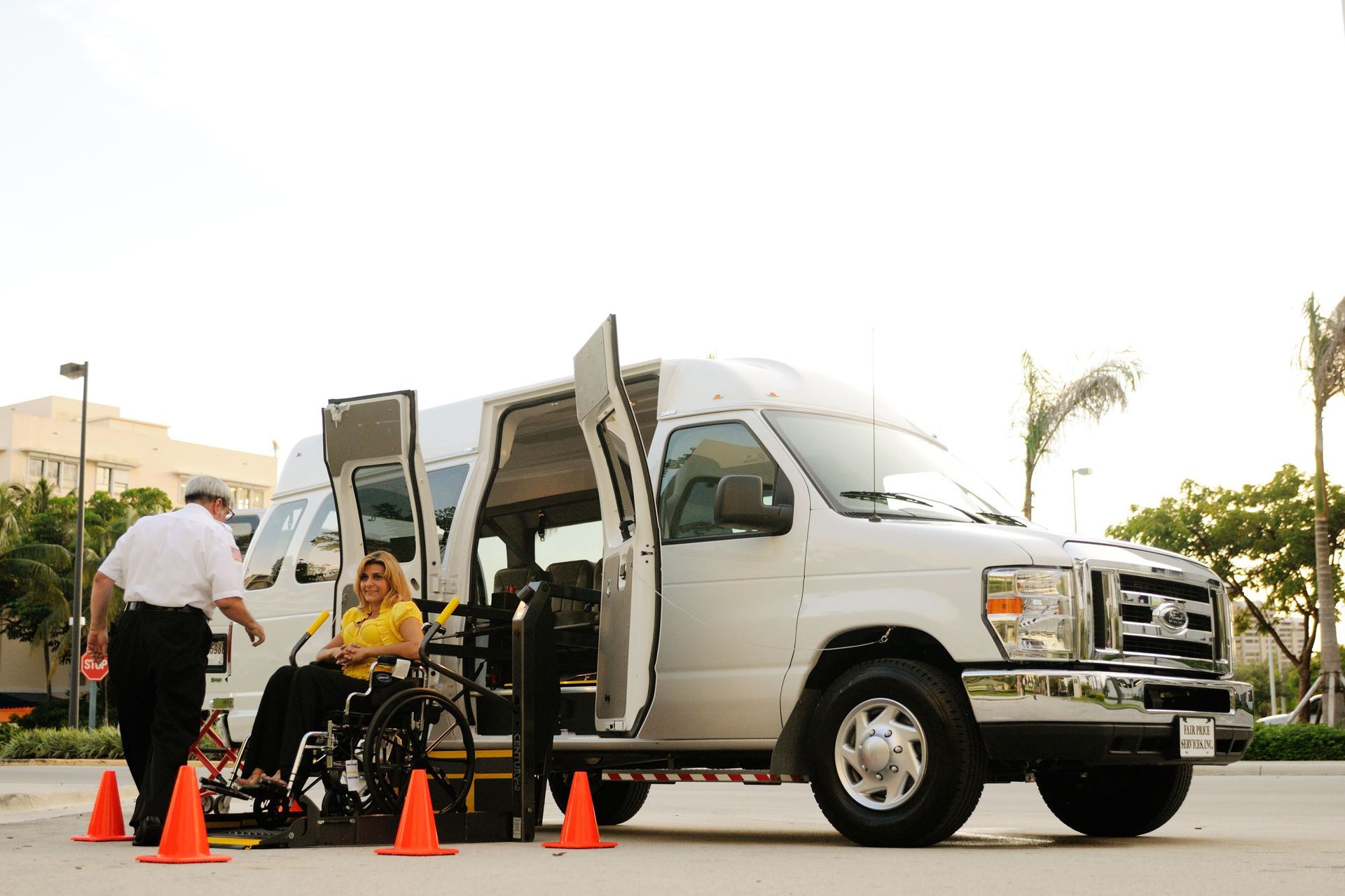 White van with open doors at a curb, cones in front, and a wheelchair user boarding with assistance