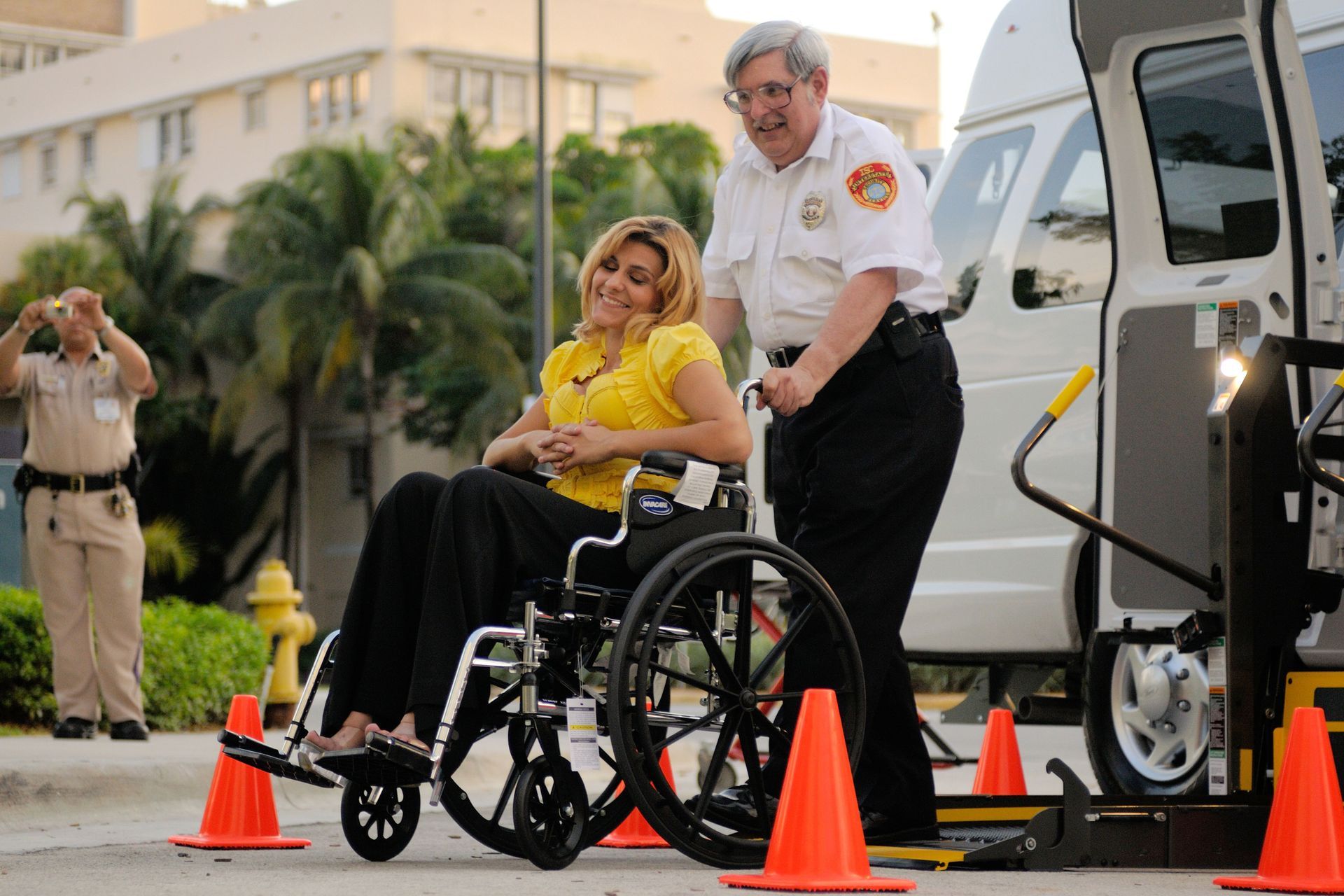 Woman in wheelchair beside a van, with an attendant and orange cones in a parking lot.