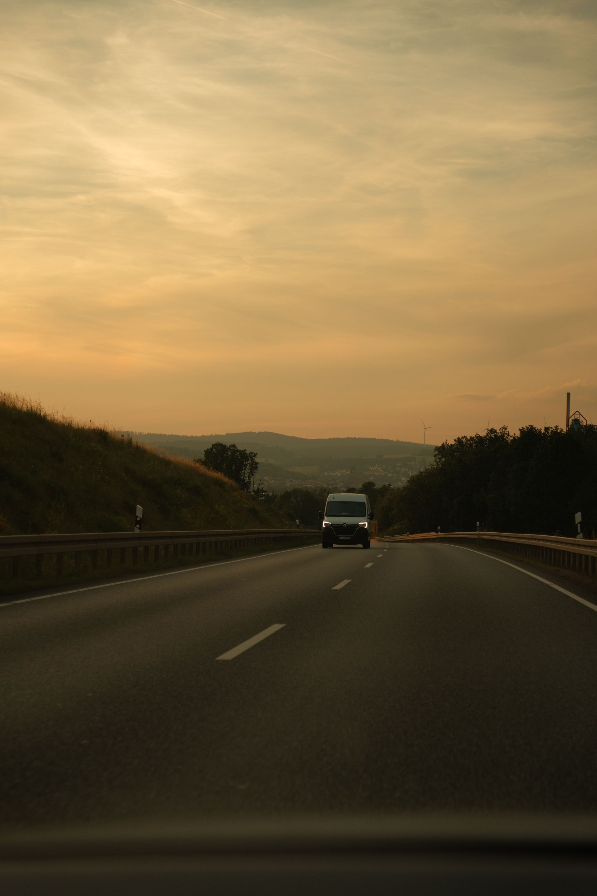 A car drives on a highway at sunset under a golden sky, with hills and roadside trees silhouetted ahead