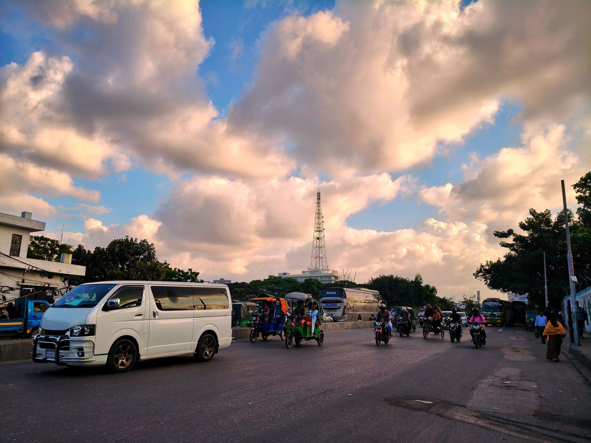 Busy street scene with motorcycles, a white van, and cloudy sunset sky near a tower