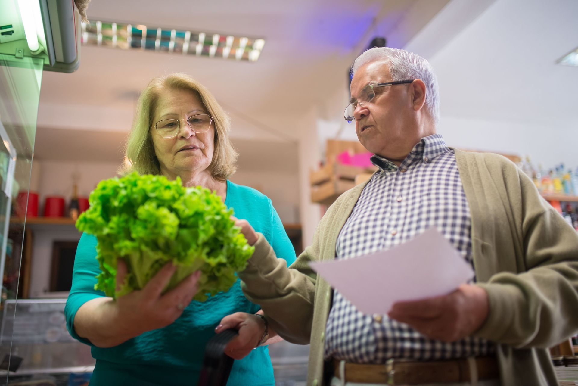 Two people in a grocery store discuss fresh lettuce while holding a receipt and paper list.