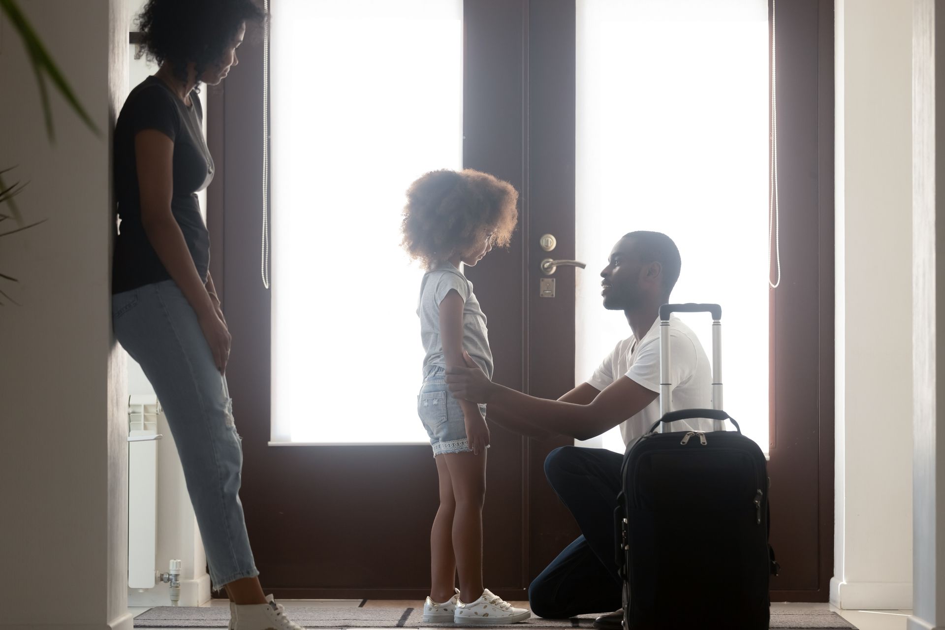 Family greeting at a front door, with a child handing something to a seated person and another adult standing nearby.